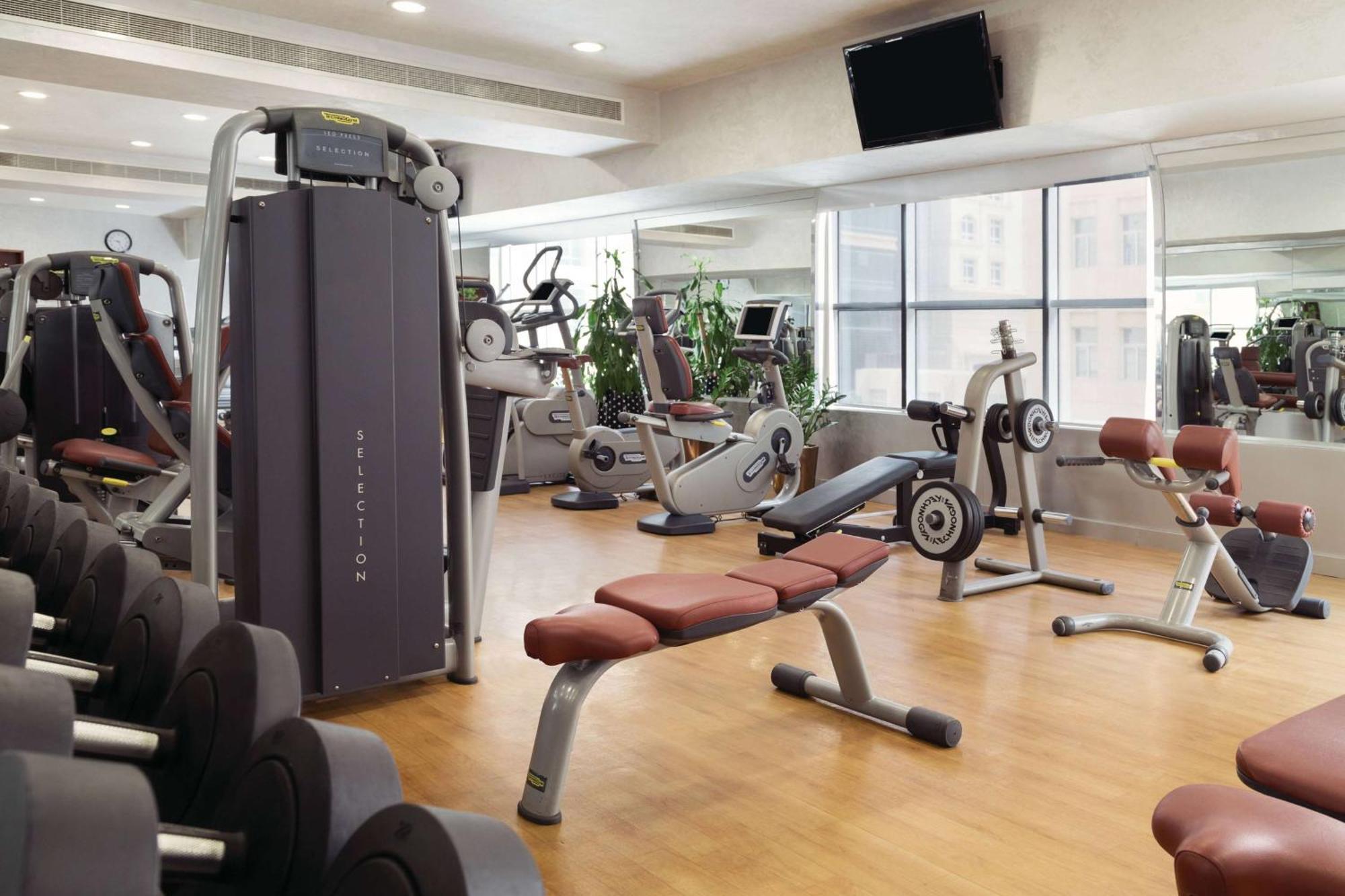 Grand Regency Doha, Trademark Collection By Wyndham Экстерьер фото The photo shows a fitness gym with various exercise equipment. There are weight machines, benches, and cardio machines such as stationary bikes and treadmills. The space has large windows allowing natural light to enter, and there are mirrors on the 